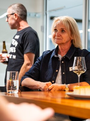 Eine Frau sitzt mit Getränk am Stehtisch, während sich Teilnehmende unterhalten.